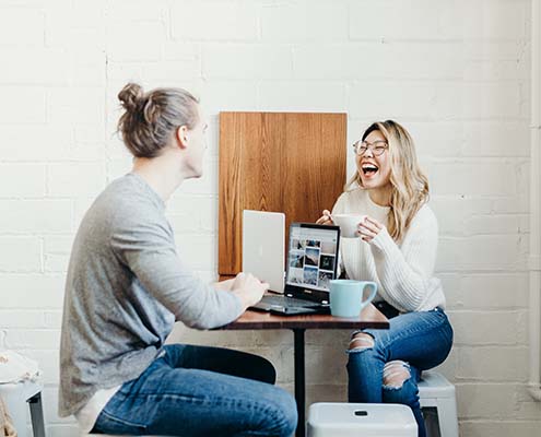A man and woman sitting at a small high-top table with their laptops and coffee mugs.